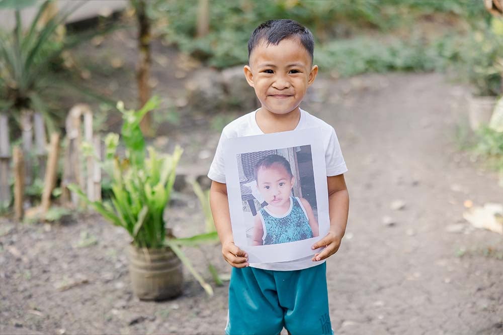 Emmanuel smiles and holds up a photo off himself from before cleft treatment